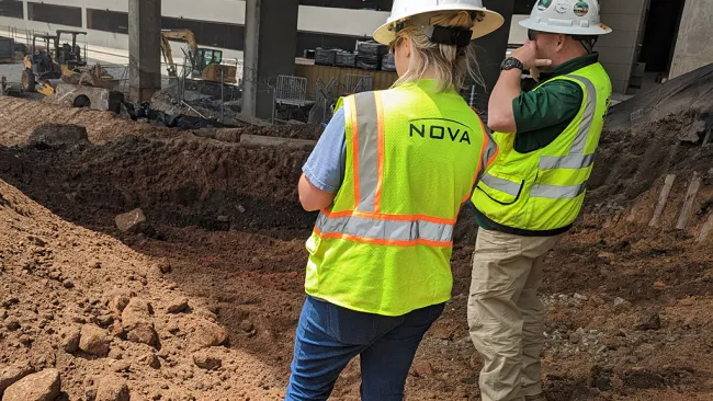 Two construction workers wearing hard hats and yellow vests standing on a dirt excavation site under a building.