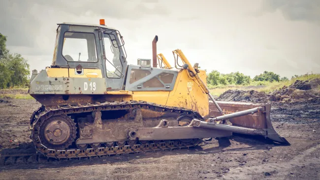 Yellow bulldozer with a rusty steel blade parked on a dirt field under a cloudy sky