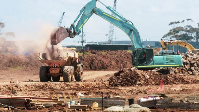 Green excavator loading dirt into a dump truck at a construction site with cranes in the background
