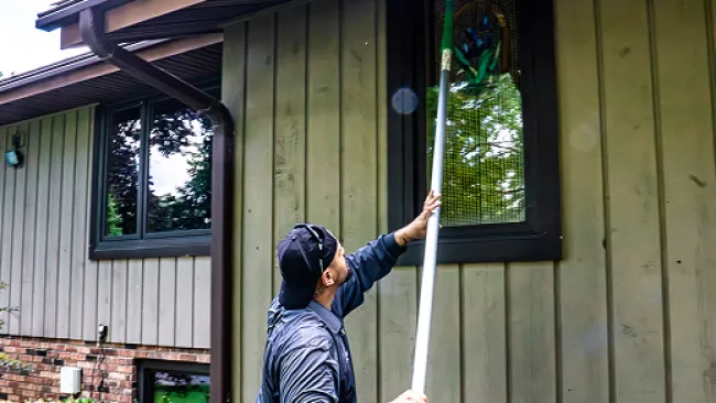 Man cleaning a high window on a house exterior using a long-handled brush during daytime.