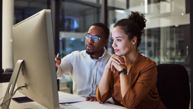 Two coworkers collaborate and discuss project details while looking at a desktop computer screen in an office.
