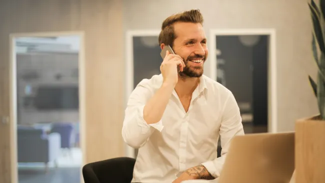 Smiling man in white shirt talking on phone while sitting at desk with laptop and coffee cup.