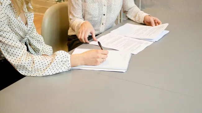 Two women reviewing and signing documents on a gray table in a professional setting