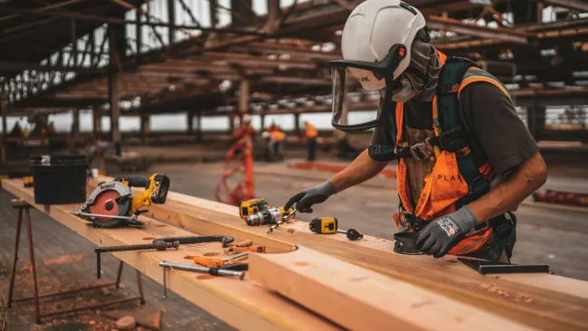 Construction worker wearing safety gear measures wood planks on a workbench inside a building frame.