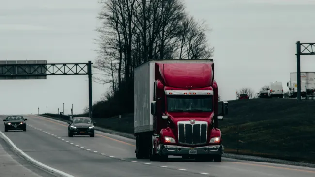 Red semi-truck driving on a highway with cars and leafless trees under cloudy sky.