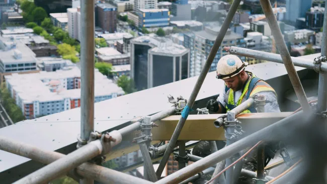 Construction worker wearing safety gear assembling scaffolding on a high-rise building with cityscape in background.