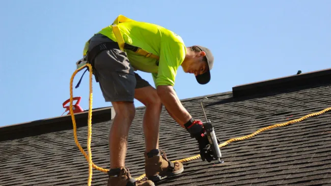 Roofer in safety gear applying sealant on shingle roof under clear blue sky during daytime.