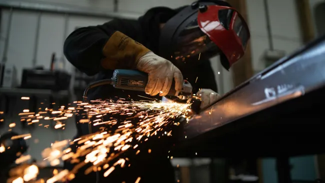 Worker wearing protective gloves and helmet using an angle grinder emitting sparks in a workshop.