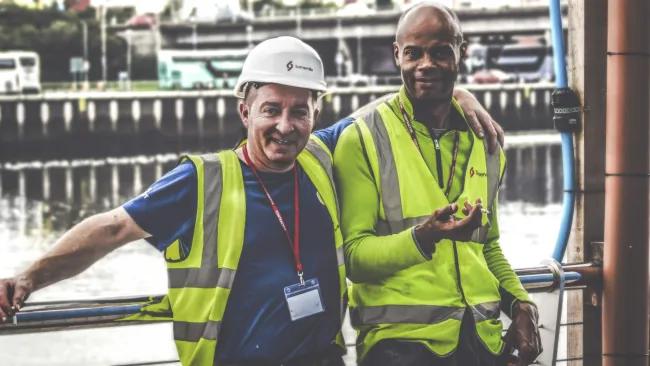 Two construction workers in safety vests and hard hats smiling by a waterfront with industrial background.