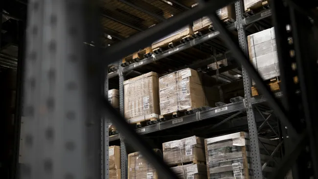 Stacked pallets of shrink-wrapped boxes stored on metal warehouse shelving racks in a large storage facility.