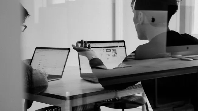 Black and white photo of two people working on laptops at a wooden table in an office setting.