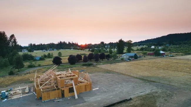 Aerial view of wooden house construction at sunset on a rural farmland with surrounding trees and distant homes.