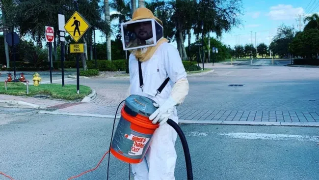 Person in protective beekeeper suit holding a homemade bee vacuum on an urban street.