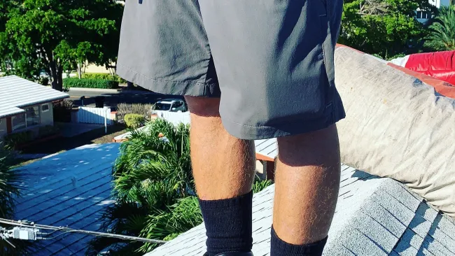 Worker wearing black boots, black socks, and gray shorts standing on a rooftop with greenery and buildings in the background.