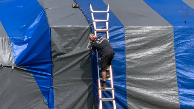Man climbing aluminum ladder to secure large blue and gray striped tarp over outdoor structure under clear sky