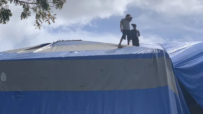 Two people standing on top of a large blue and gray tarp-covered structure under cloudy sky near a tree.