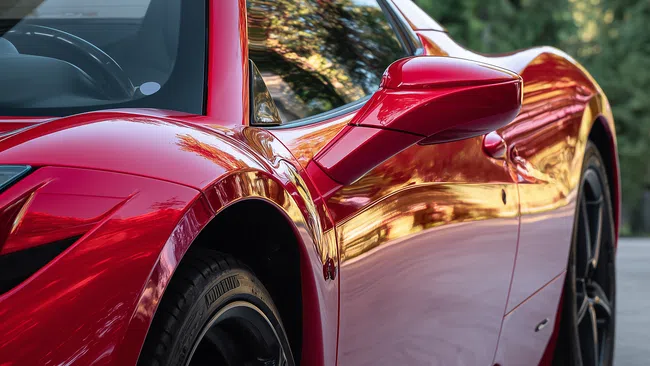 Close-up of a shiny red sports car side with sleek curves, black alloy wheel, and reflective windows.