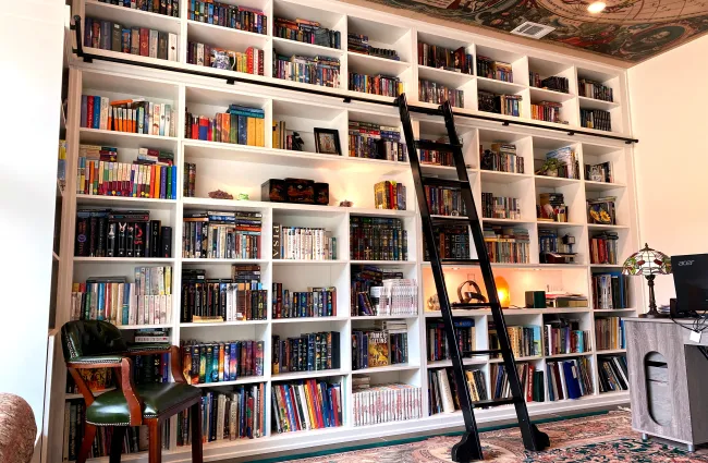 Large white bookshelf filled with colorful books, rolling ladder, green leather chair, and patterned rug.