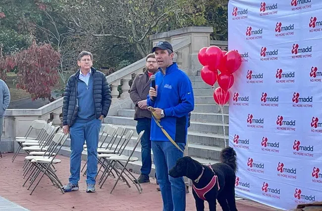 Man speaking at outdoor event with dog, red balloons, and MADD No More Victims backdrop and seated audience.