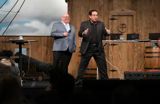 Two men on stage speaking at an event with wooden barrels and laptops in the background