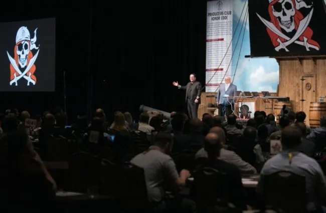 Audience watches speakers presenting on stage with pirate skull and crossbones on large banners at conference.