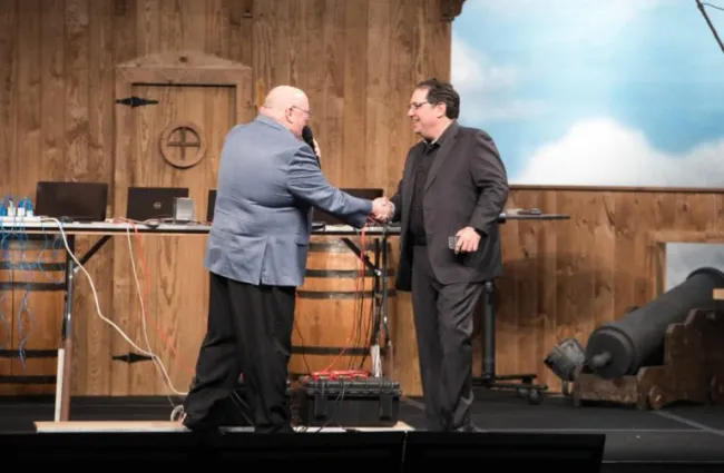 Two men in suits shaking hands on stage with microphones in a wooden rustic setting with tech equipment.