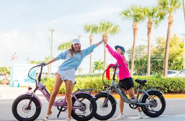 Two women high-fiving while standing next to electric bikes on a sunny street with palm trees.