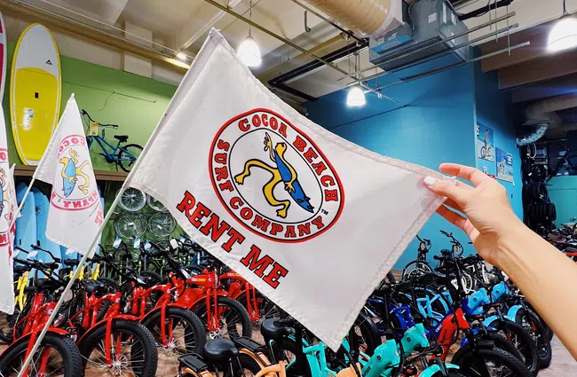 Hand holding a Cocoa Beach Surf Company flag in front of colorful rental bikes inside a shop with surfboards.