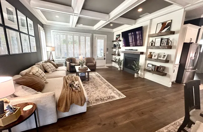 Modern living room with beige sofa, brown armchairs, fireplace, wooden floor, and large windows with shutters.