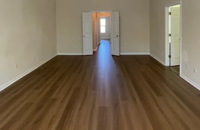Empty room with wood flooring, white walls, ceiling fan with light, and a doorway leading to a bright hallway.