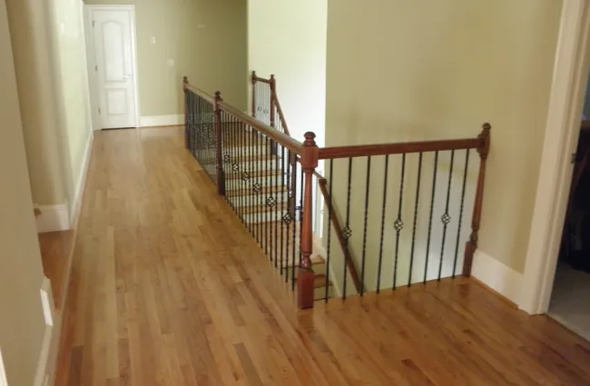 Wooden floor hallway with black metal and wood railing overlooking staircase and beige walls.