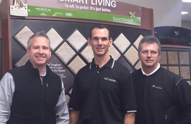 Three men in black shirts pose in front of a carpet display board at a flooring store.