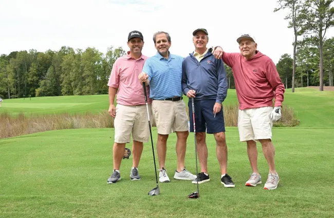 Four men standing on a golf course, smiling, holding golf clubs with trees in the background on a clear day