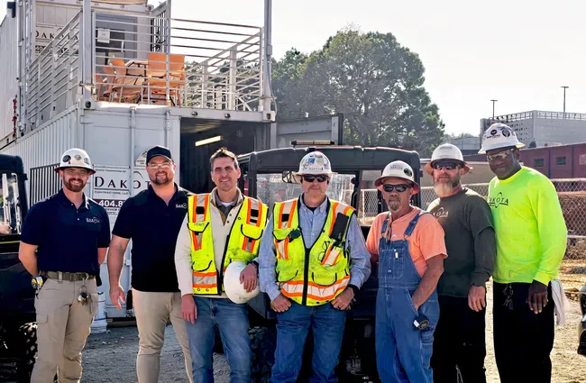 Seven construction workers wearing safety gear pose at a job site with equipment and containers in the background