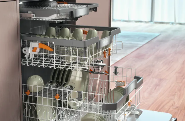 Open dishwasher loaded with clean dishes, cups, and bowls in a modern kitchen with wooden floor.