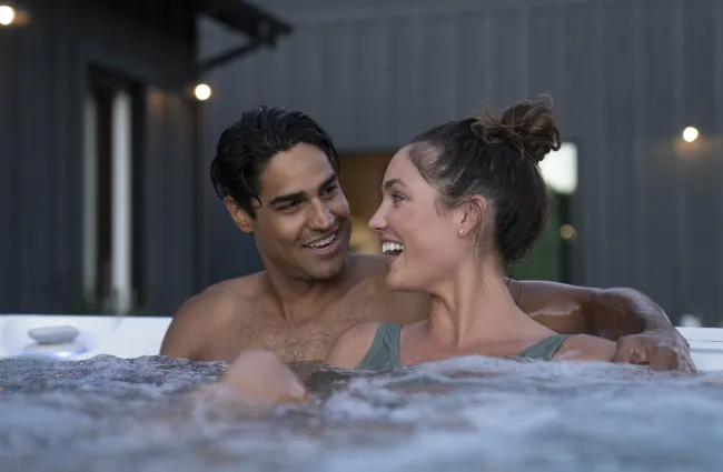 Smiling couple relaxing and talking in an outdoor hot tub with modern house background at dusk.