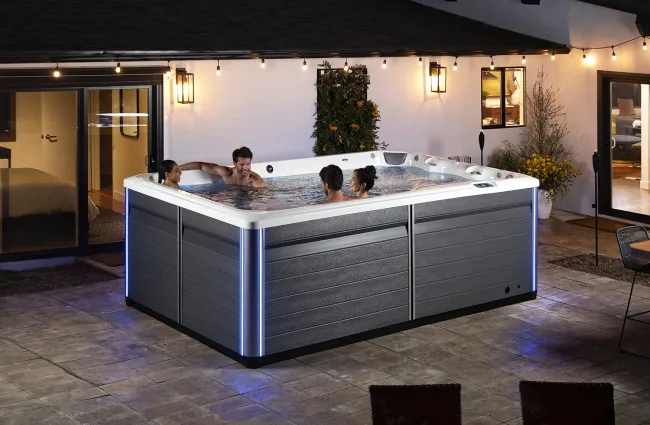 Four people relaxing in a modern outdoor hot tub with blue LED lights on a patio at night.