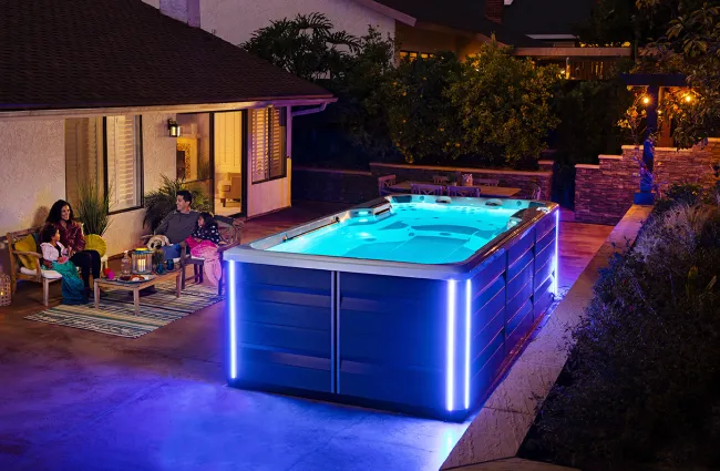Modern backyard with illuminated blue swim spa and family relaxing together at night on patio seating.
