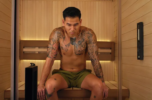 Tattooed man wearing green shorts sitting in a wooden sauna room with focused expression