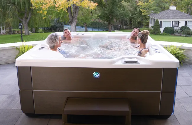 Four people enjoying a brown outdoor hot tub on a patio surrounded by trees and a house in the background.
