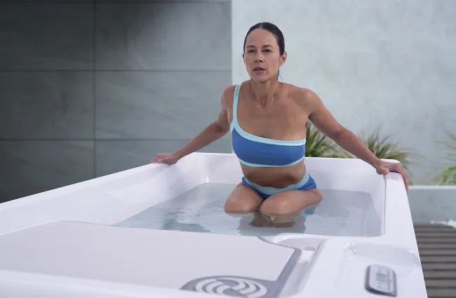 Woman in blue swimwear stepping into a modern outdoor hot tub with water and a serene background.