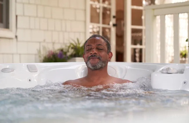 Man relaxing with eyes closed in a bubbling outdoor hot tub on a bright day.
