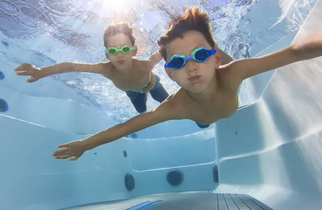 Two boys wearing goggles swimming underwater in a sunlit clear pool with arms outstretched.