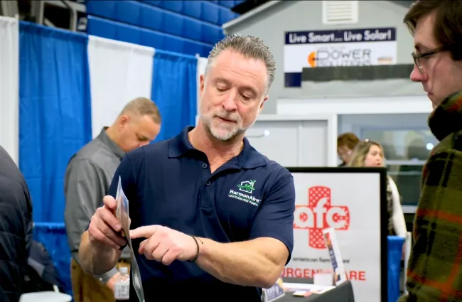 Man explaining product details to visitor at an indoor trade show booth with informational displays.