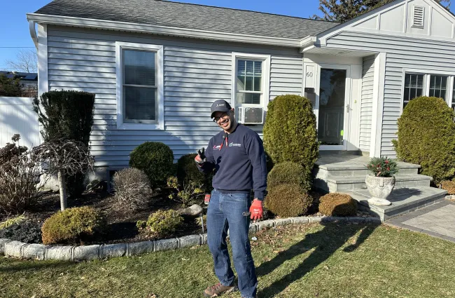 Smiling landscaper giving thumbs up in front of a well-maintained suburban house with garden beds.