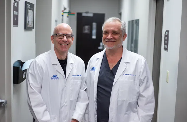Two doctors in white lab coats smiling in a medical office hallway.