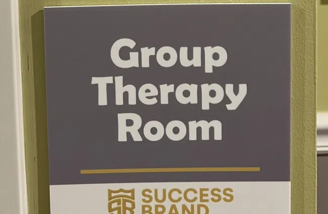 Signs for classroom 103 and group therapy room on a yellow wall with educational and success branding logos.