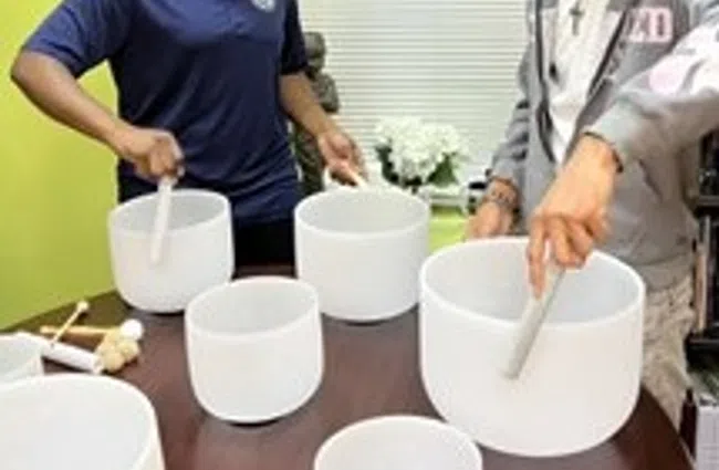 Two people playing white crystal singing bowls on a wooden table in a bright room with green walls.