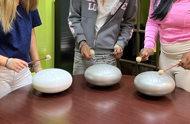 Three people joyfully playing handpans around a round wooden table in a bright room.