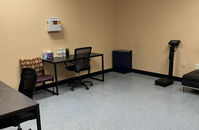 Minimalist office room with beige walls, desks, chairs, water bottles, scale, and medical equipment.